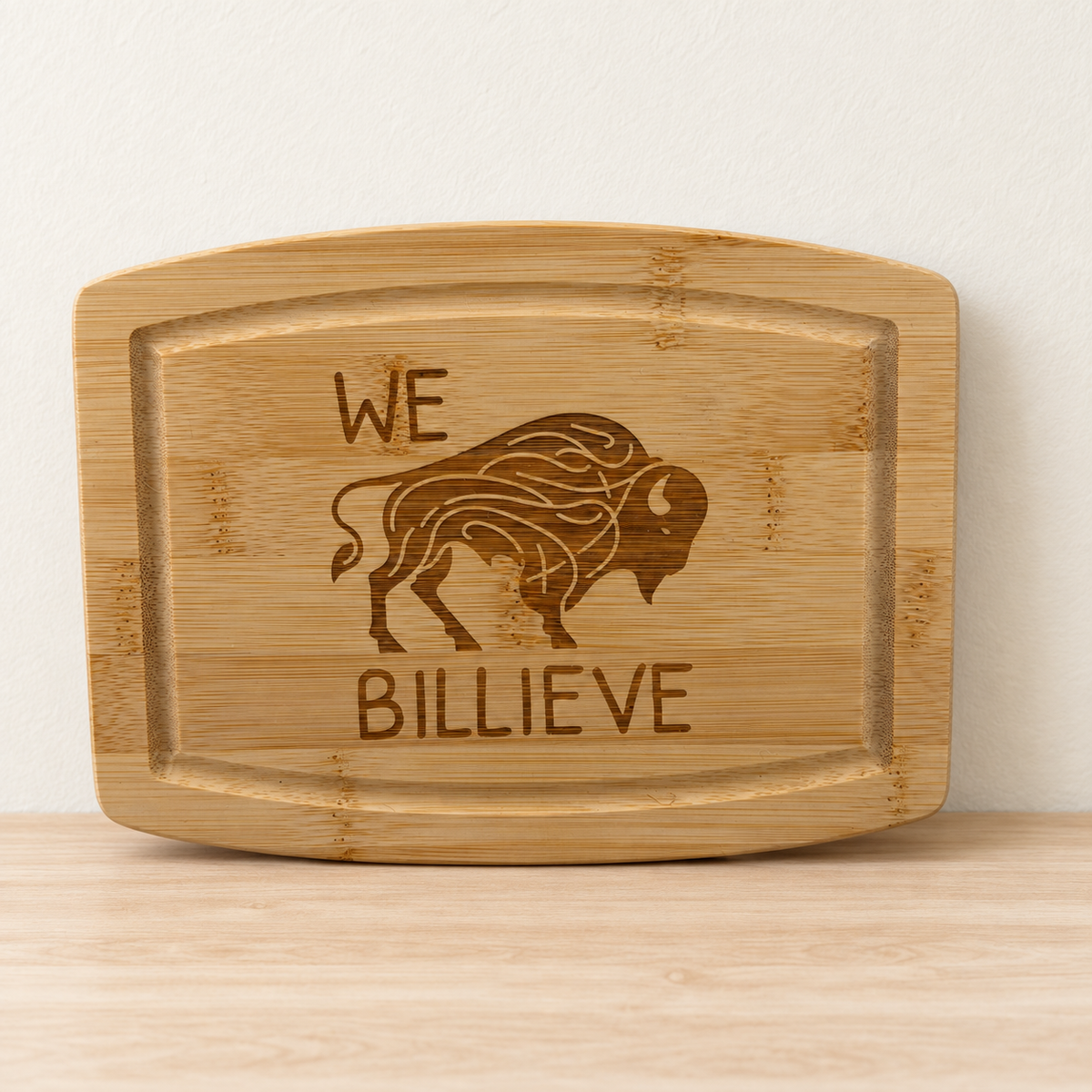 Bamboo cutting board with a buffalo engraving and the words We Billieve, displayed against a cream wall on a pale wood counter.