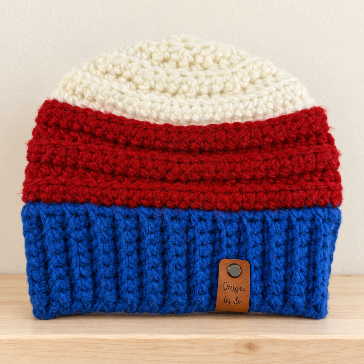 Crocheted beanie with cream, red and royal blue bands and a brown Designs by Lo tag, resting on a pale wooden shelf.