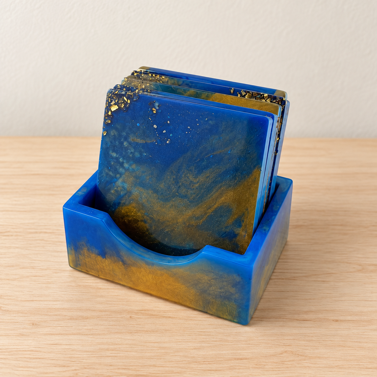 Blue and gold square coasters in a matching holder with a curved front opening, displayed on a pale wood surface.