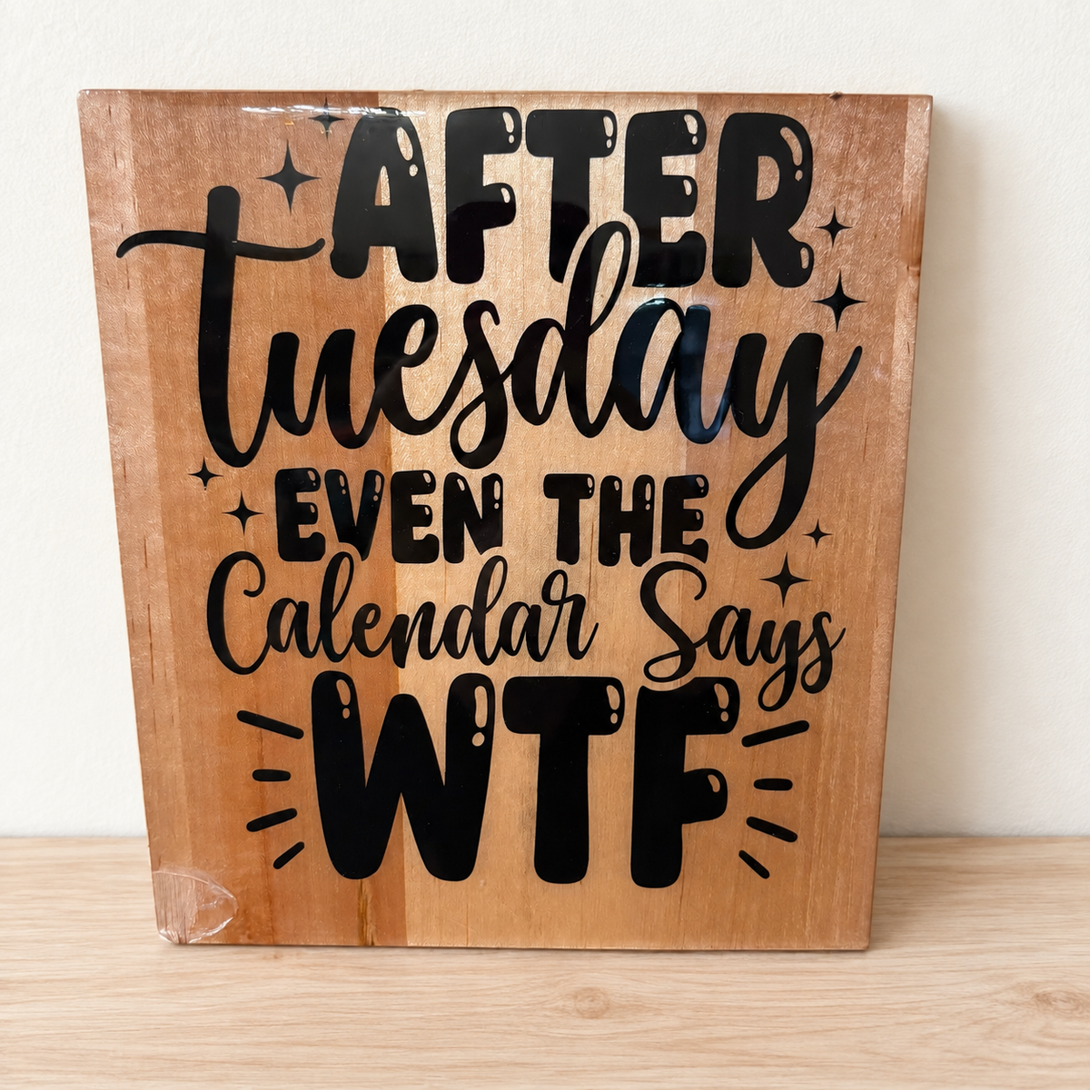 Wood-tone After Tuesday sign with bold black lettering and sparkle details, displayed against an ivory wall on pale wood.