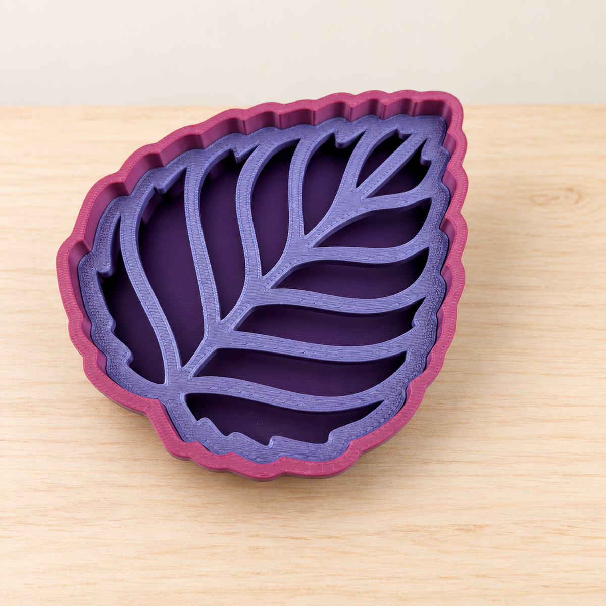 Leaf-shaped purple soap dish with a magenta scalloped rim and leaf-vein drainage insert, shown empty on pale wood.